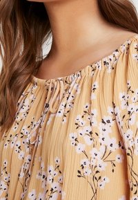Nærbillede af en kvindes skulder og bryst iført en gul blomstret bluse med rynket halsudskæring og små bånddetaljer.