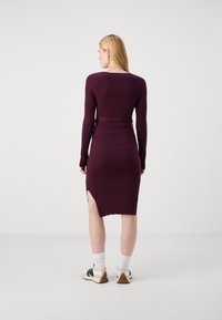 Long burgundy ribbed dress with long sleeves, side slit, and fitted silhouette. Worn with white socks and sneakers. Plain background.
