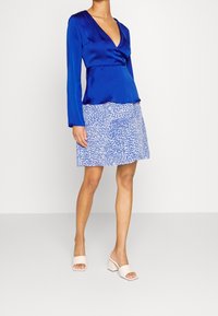 Top blu satinato a maniche lunghe, modello incrociato, abbinato a una gonna blu a fantasia. Calzature: mules con tacco a blocco color crema, design aperto davanti.