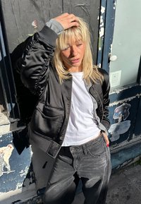 Fekete selyem szatén bomber dzseki, begyűrt ujjakkal, szürke részletekkel, fehér pólóval és kifakult fekete farmerrel, városi környezetben, kék fallal.