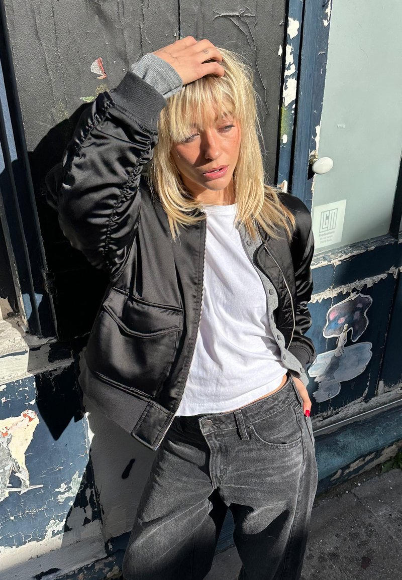 Fekete selyem szatén bomber dzseki, begyűrt ujjakkal, szürke részletekkel, fehér pólóval és kifakult fekete farmerrel, városi környezetben, kék fallal.