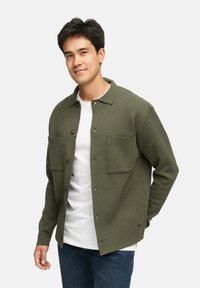 Grön, texturerad skjortjacka med knäppning framtill, två bröstfickor och krage. Bärs över en vit t-shirt, matchad med blå jeans.