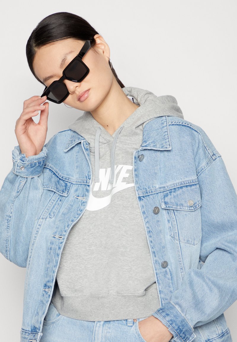 Světle modrá džínsová bunda přeložená přes šedou mikinu s bílým logem. Černé hranaté sluneční brýle, hladká texturer, ležérní střih.