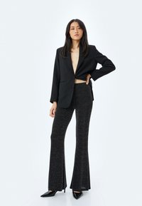 Zwarte blazer met een diepe V-hals, gecombineerd met uitlopende, getextureerde zwarte broeken. Model draagt zwarte spitse hakken. Eenvoudige achtergrond.