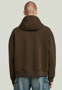Bruine hoodie, oversized ontwerp, zachte textuur, ribgebreide mouw- en zoomopening, achteraanzicht dat de capuchon en minimalistische details benadrukt.