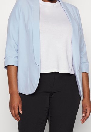 Vrouw die een lichtblauw colbert draagt over een wit overhemd en zwarte broek met opgerolde mouwen tegen een effen achtergrond.