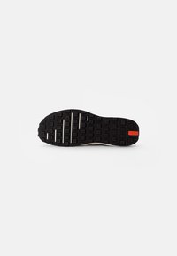 Semelle de chaussure avec des crampons noirs en forme de carrés et de petits accents rectangulaires beiges, avec un petit détail rouge près du talon.