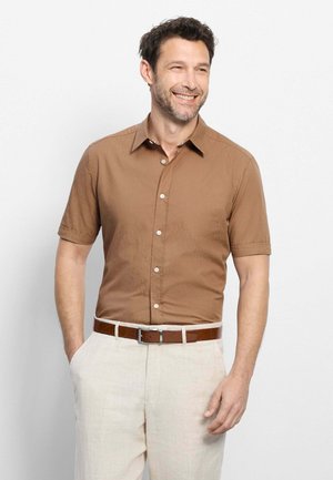 Homme souriant portant une chemise marron à manches courtes boutonnée, un pantalon beige et une ceinture marron, debout avec une main dans la poche.