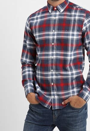 Mand iført en ternet skjorte i rød, hvid og blå med hænderne i lommerne og mørkeblå jeans.