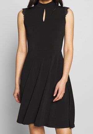 Femme portant une robe noire sans manches avec des manches courtes en dentelle, un col haut et une petite découpe en forme de trou de serrure au niveau de la poitrine.