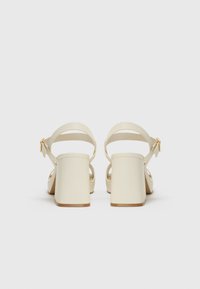 Sandales à talons beiges avec un talon carré épais, de fines brides autour de la cheville et un détail de boucle, fabriquées en matériau synthétique lisse.