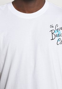 Weiße Baumwoll-T-Shirt mit rundem Ausschnitt. Auf der linken Brust befindet sich ein Grafikdruck in Schwarz und Türkis mit der Aufschrift "the Great Beach Club".