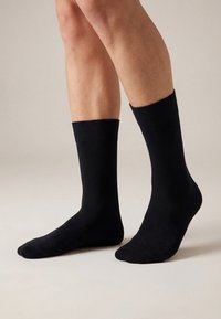 Dvije noge u običnim crnim čarapama duljine do sredine lista, stoje na neutralnoj bež pozadini; jedna noga je ravno na tlu, a druga je lagano podignuta.