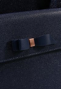 Ciemnoniebieska torebka skórzana o teksturze, z błyszczącym wykończeniem, w kształcie prostokąta, z akcentem w postaci kokardy i mosiężnym detalem.