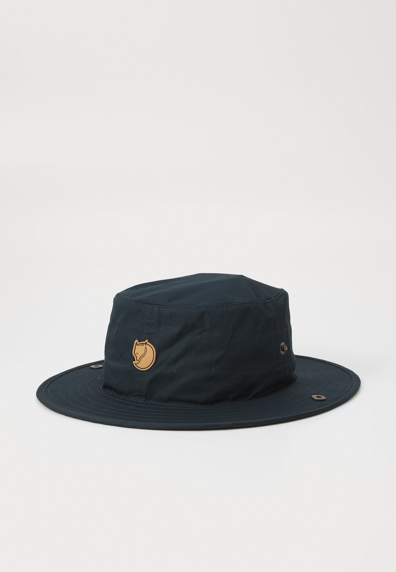 Marineblauer Bucket Hat aus leichtem Stoff mit runder Krone, breiter Krempe und einem braunen Logo-Patch auf der Vorderseite. Verfügt über Belüftungsösen.