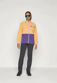 Casaco de zíper laranja claro e roxo com gola alta, bolsos frontais e design em bloco de cores. Usado com calças pretas e botas.