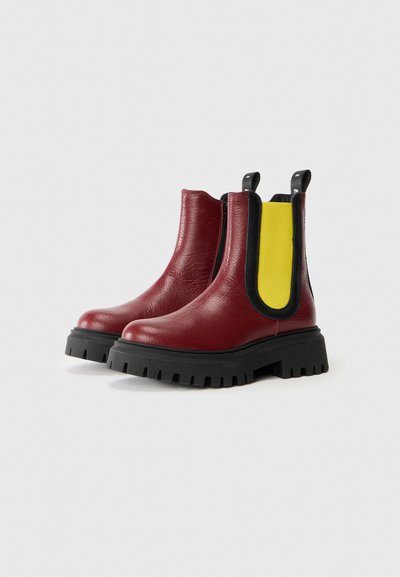 Röda läderankelboots med kraftig svart sula, med en gul elastisk detalj och svarta sidopaneler. Texturerad yta och dragflikar.
