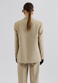 Beige tailored blazer with a textured finish, wide lapels, and four-button cuffs, paired with matching trousers. Black gloves and oversized earrings.