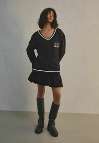 Černá oversized mikina s V-výstřihem a bílými pruhy, spárovaná s černou sukní s volány a černými kozačkami ke kolenům, s retro textem.