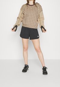 Hnědý pulovr s geometrickým vzorem, doplněný černými sportovními šortkami. Model drží odporové gumy a má na sobě černé tenisky.