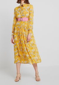 Robe midi à imprimé floral jaune avec des manches longues et un col noué, cintrée à la taille avec une large ceinture rose. Sandales beiges à brides.