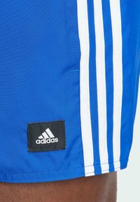 Pantaloni sportivi blu con strisce bianche, texture del tessuto e una patch con il logo Adidas sull'orlo. Progettati per l'abbigliamento sportivo.