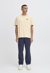 Lys gul kortærmet T-shirt med sort solgrafik, parret med marineblå bukser og beige sneakers, stående mod en ensfarvet baggrund.