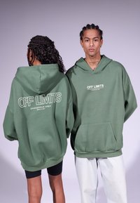 Grønne overdrevne hettegensere med frontlomme, hvit tekst "OFF LIMITS DANGEROUS AREA" på bak- og framsiden. Modellen har på seg svarte shorts og lyse jeans.