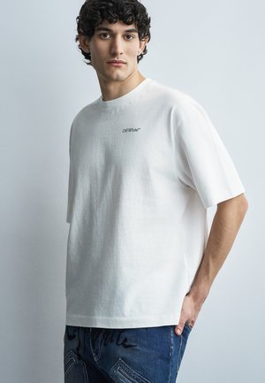 Młody mężczyzna z kręconymi włosami, ubrany w białą koszulkę Off-White i niebieskie dżinsy, stoi na tle jednolitego jasnoszarego tła.