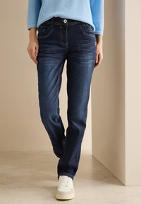 Jeans de mezclilla azul oscuro con un corte recto, que presentan un diseño de cinco bolsillos, costuras en contraste y un cierre de un solo botón. Zapatillas blancas visibles.