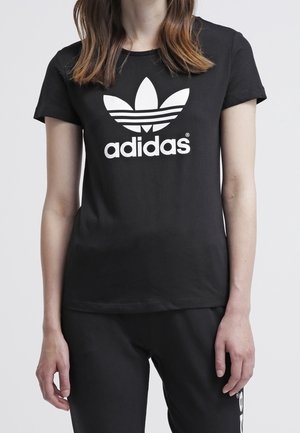 Osoba ubrana w czarną koszulkę Adidas z dużym białym logo Adidas i czarne spodnie na jednolitym jasnym tle.