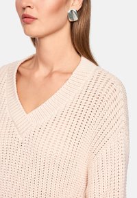 Light pink, knitted sweater with a V-neckline, showcasing textured patterns; paired with square, metallic earrings with subtle sheen.