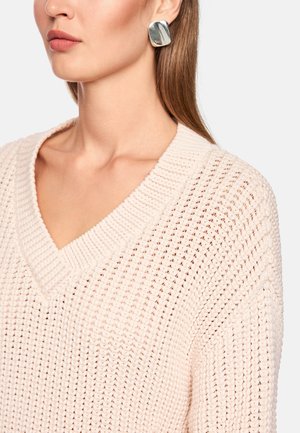 Light pink, knitted sweater with a V-neckline, showcasing textured patterns; paired with square, metallic earrings with subtle sheen.