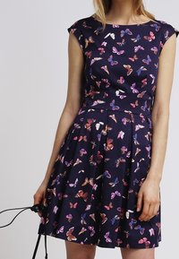 Kvinna klädd i en marinblå klänning med färgglada fjärilstryck, åtsittande midja, korta ärmar, hållande en svart rem eller snöre i vänster hand.