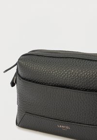 Fekete bőr erszény texturált felülettel, felül cipzáras záródással és egy finom logóval az elején. Téglalap alakú.