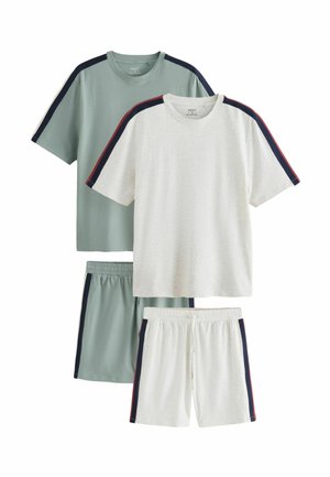 Un homme pieds nus se tient dans une chambre, portant un short bleu-gris assorti et un T-shirt avec des rayures sombres sur les épaules et les côtés.