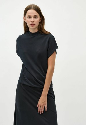 Black, draped, short-sleeve top with a high neckline, made of soft fabric. Woman wearing it with a matching black skirt, standing against a white background.