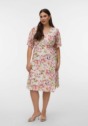 Kobieta ubrana w sukienkę kopertową z kwiatowym wzorem do kolan, z krótkimi rękawami, oraz beżowe sandały na obcasie, stojąca na tle białego, gładkiego tła.