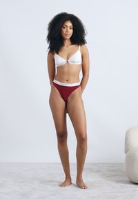 Balta krūšturis ar plāniem siksniņām un dekoratīvu pogu, kopā ar bordo augsto vidukļu biksītēm ar zīmola jostasvīru. Mīksts fons.