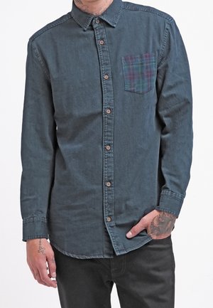 Hombre con camisa azul oscuro abotonada con un bolsillo a cuadros en el pecho y pantalones negros, mostrando tatuajes en sus manos y muñecas.