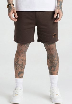Mann iført brune shorts med logo på venstre ben, hvite crew-sokker, hvite sneakers og tatoverte legger og hender.