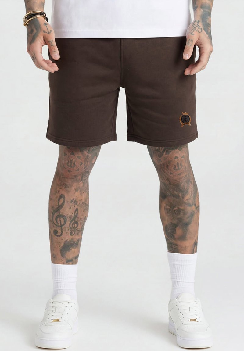 Homme portant un short marron avec un logo sur la jambe gauche, des chaussettes blanches montantes, des baskets blanches, avec les jambes et les mains tatouées.