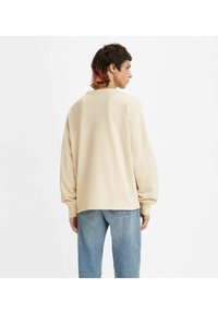 Sudadera de color beige claro, de corte oversized, con mangas largas, escote redondo y dobladillos ajustados, combinada con jeans rectos de color azul claro.