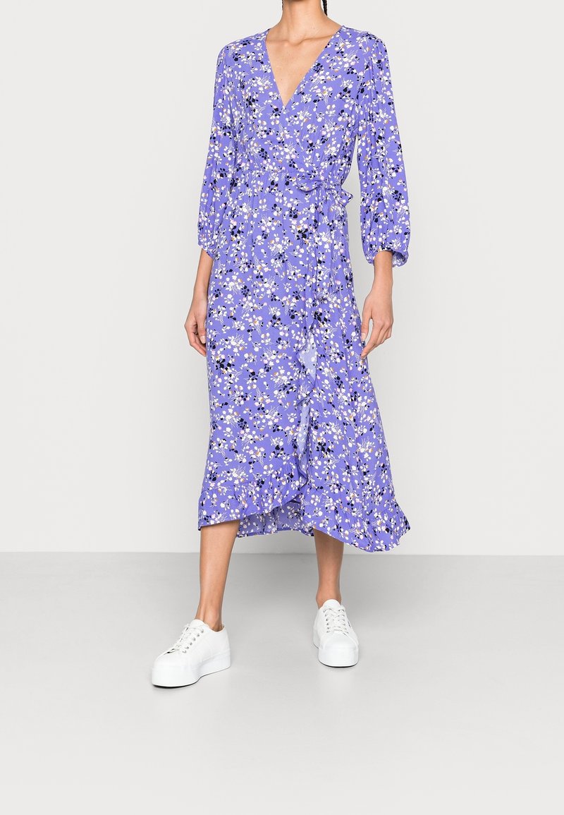 Violetti kukkakuosiset kietaisumekko, jossa on kolmivartiset puffihihat, röyhelöhelma ja valkoiset kangaskengät. Mekossa on syvä V-pääntie ja kukkakuosi.