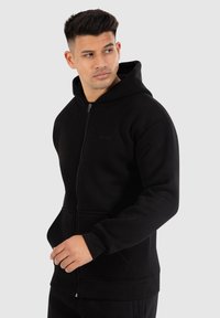 Zwarte rits hoodie van zachte stof met een voorkant zak, verstelbare capuchon en geribde mouwen en zoom. Bevat een klein logo op de borst.