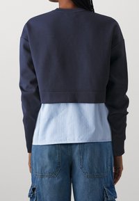 Sweat-shirt court marine à manches longues porté sur une chemise à rayures bleu clair. Caractérisé par une texture douce et un design superposé.