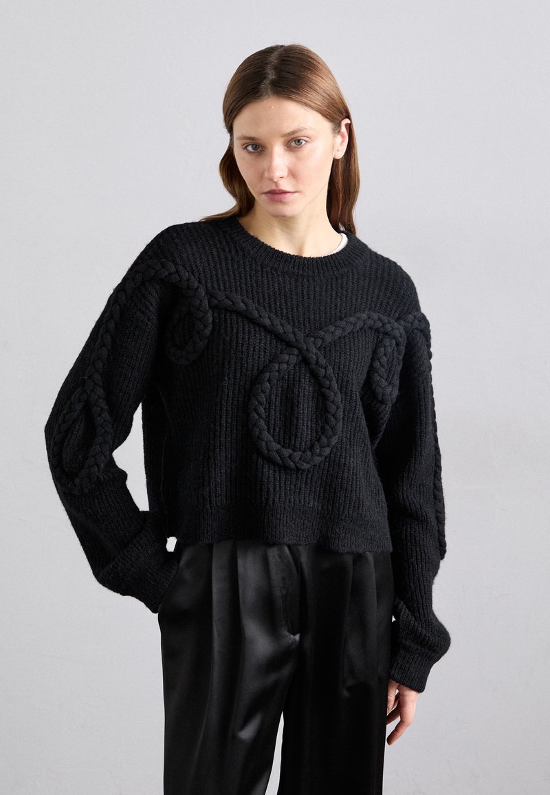 Schwarzer gestrickter Pullover mit geflochtenem Muster über der Brust und an den Ärmeln; verkürztes Design mit gerippten Bündchen und Saum, kombiniert mit schwarzen Hosen.