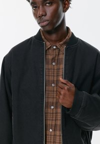 Crna bomber jakna s rebrastim manžetama i ovratnikom, sadrži srebrni patentni zatvarač i smeđu kariranu košulju ispod s dugmadi na preklop.