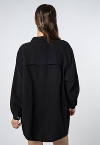 Black oversized shirt in textured fabric with long sleeves, collared design, and a back yoke detail, featuring buttoned cuffs and side slits.