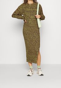 Outfit lavorato a maglia in tonalità di verde e marrone, caratterizzato da un top a maniche lunghe con texture e una gonna midi abbinata con uno spacco laterale. Indossato con sneakers.